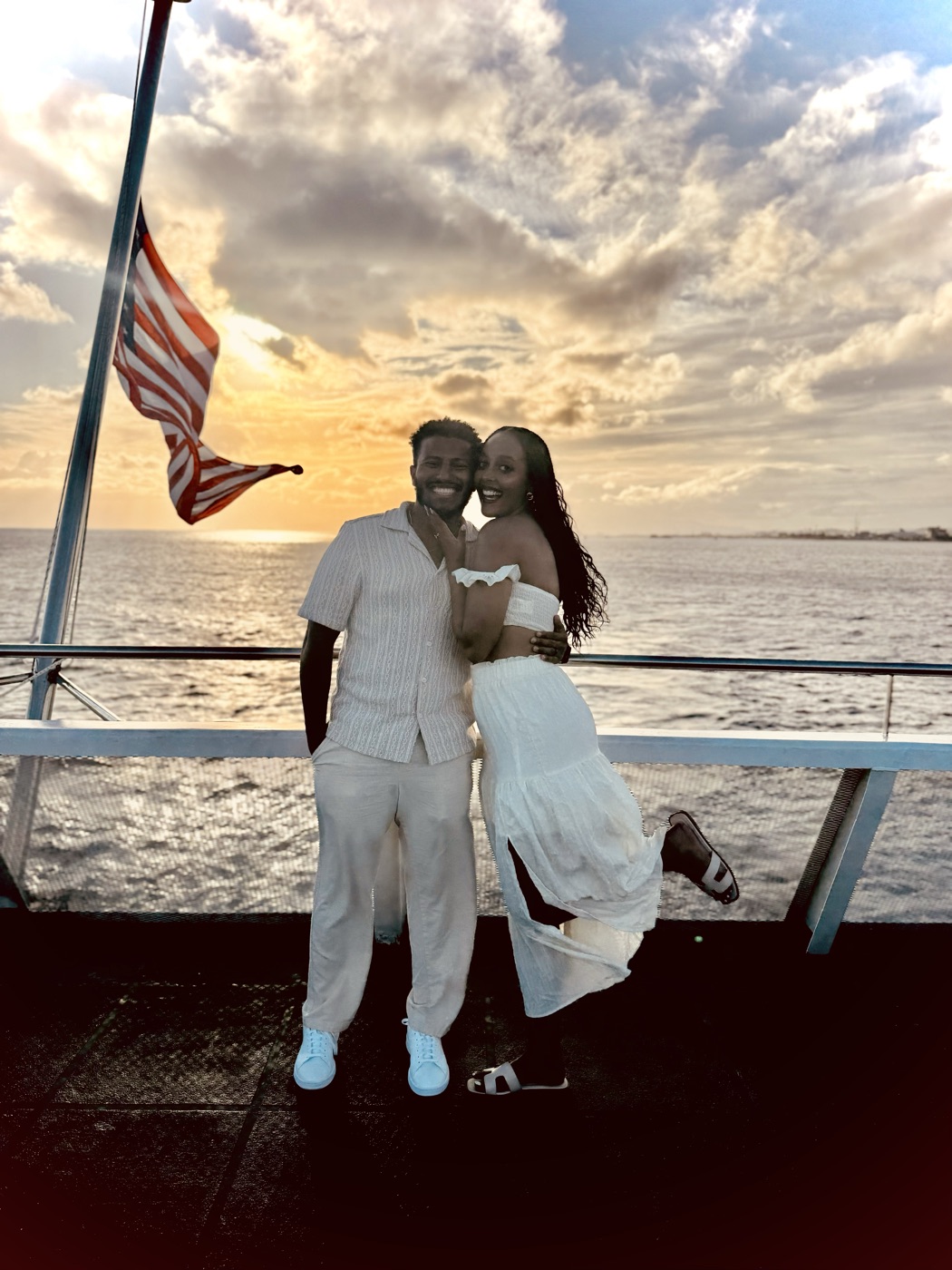 Edosa laughing with their wife on a boat deck at sunset, an American flag waving behind them