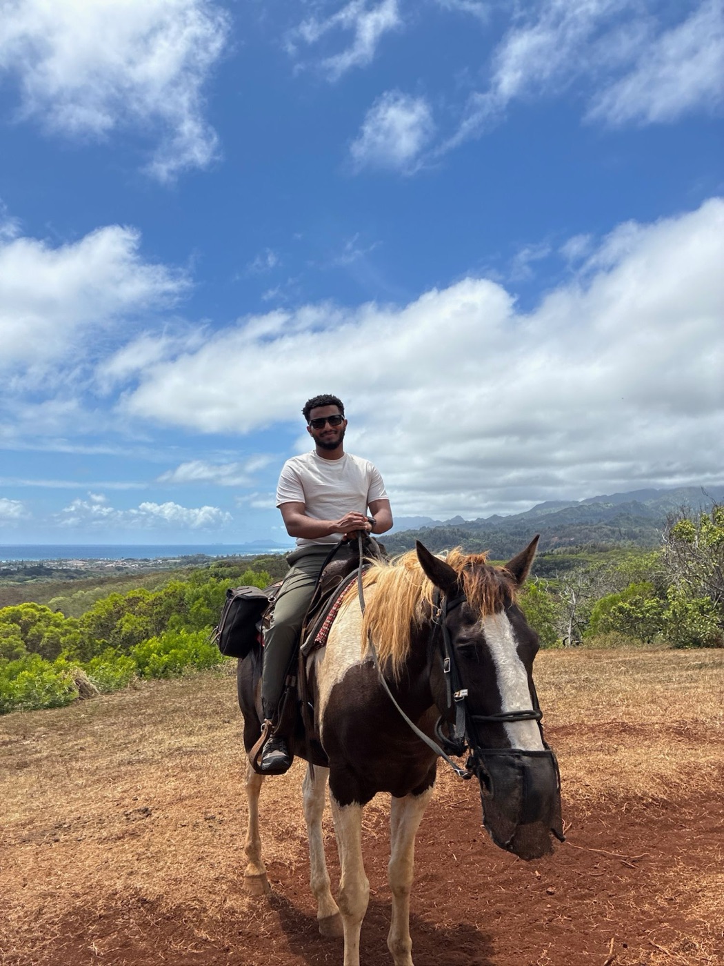 Edosa smiling in sunglasses on a painted horse, green hills and the ocean in the background