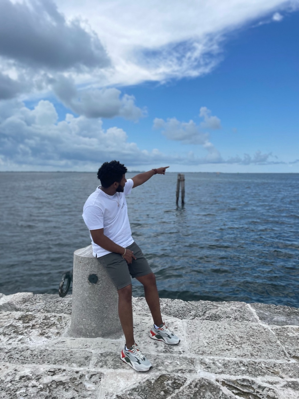Edosa sitting on a stone bollard on a pier, pointing out across the water under a big blue sky