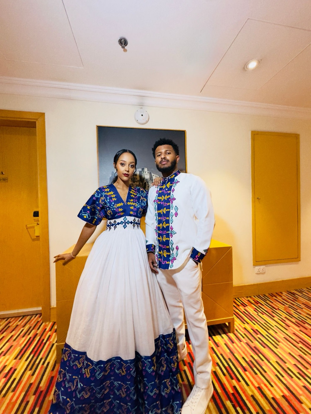 Edosa and their wife in matching white outfits with blue embroidered patterns, posing in a hotel hallway