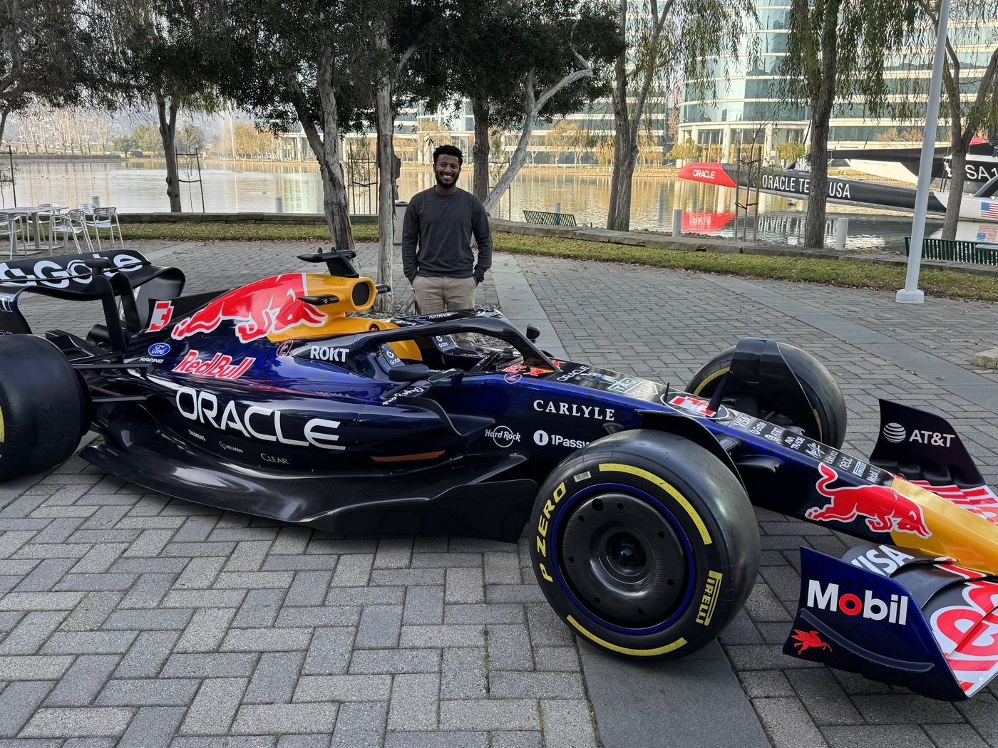 Edosa standing behind a Red Bull Racing Formula 1 car with Oracle branding, parked by the lake at Oracle's campus