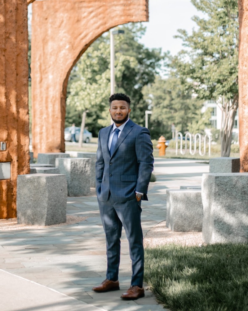 Edosa Leta standing among clay-colored arches on a sunny campus path